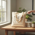 The Witches' Quest beige canvas tote bag with black floral line art and Grow positive thoughts text on a wooden desk.