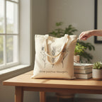 The Witches' Quest beige canvas tote bag with black floral line art and Grow positive thoughts text on a wooden desk.