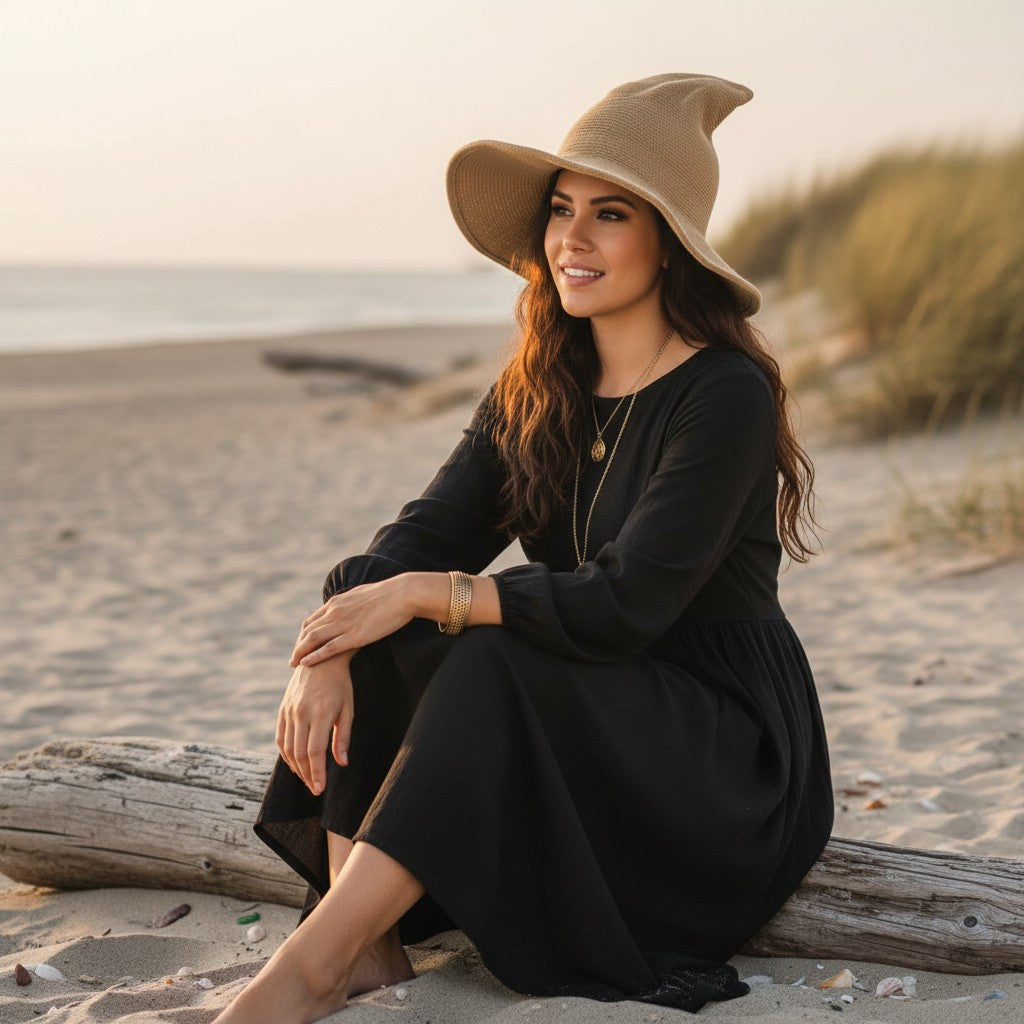 The Witches' Quest beige knit witch hat with a wide brim worn by a woman in a black dress on a sandy beach at sunset.