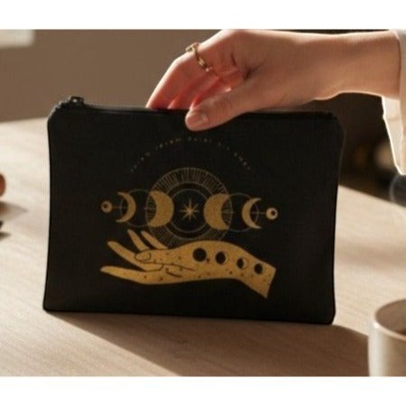 The Witches' Quest black canvas zipper pouch featuring a gold moon phase and hand design held by a hand