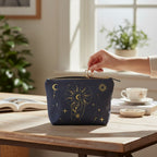 The Witches' Quest black canvas clutch with gold celestial moon and sun print, held on a wooden table.