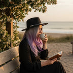 The Witches' Quest black wide brim fedora with floral embroidery worn by a woman reading on a beach bench at sunset.