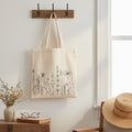 The Witches' Quest beige canvas tote bag with black wildflower line art hanging on a wall hook.