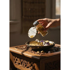 The Witches' Quest Ash & Sea Gold Dust ritual powder being sprinkled from a glass shaker over a bowl of gold coins.