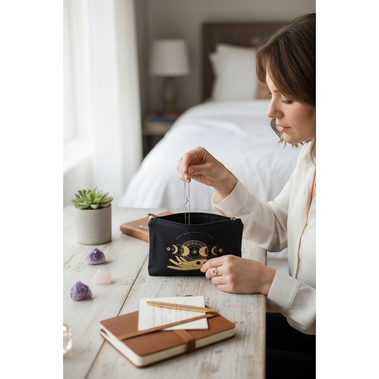 The Witches' Quest black canvas zipper pouch featuring a gold moon phase and hand design held by a woman at a desk.
