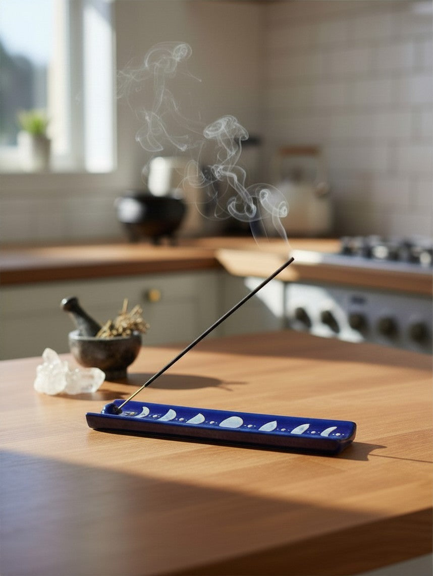 The Witches' Quest midnight blue ceramic incense holder with moon phase design, holding a smoking incense stick.