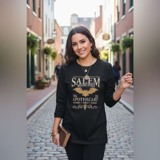 The Witches' Quest black long sleeve shirt with a gothic Salem Apothecary graphic worn by a smiling woman outdoors.