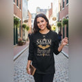 A woman wearing The Witches' Quest black long-sleeve crewneck shirt with a gold Salem Apothecary bat graphic on a street.