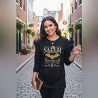 A woman wearing The Witches' Quest black long-sleeve crewneck shirt with a gold Salem Apothecary bat graphic on a street.