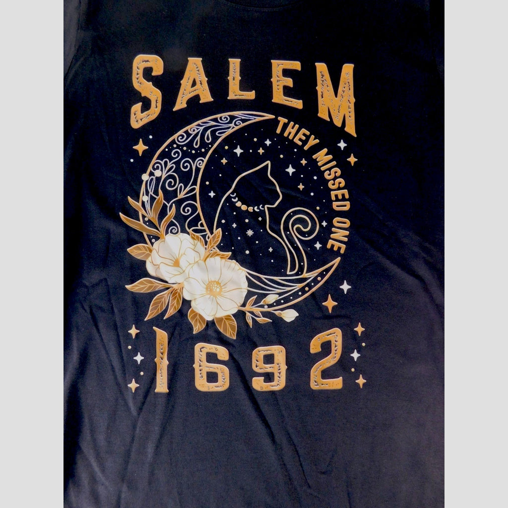 The Witches' Quest black cotton unisex t-shirt featuring a Salem 1692 graphic with a celestial cat and floral design.