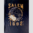 The Witches' Quest black cotton unisex t-shirt featuring a Salem 1692 graphic with a celestial cat and floral design.