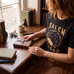 The Witches' Quest black cotton unisex t-shirt featuring a Salem 1692 celestial cat and floral graphic print.