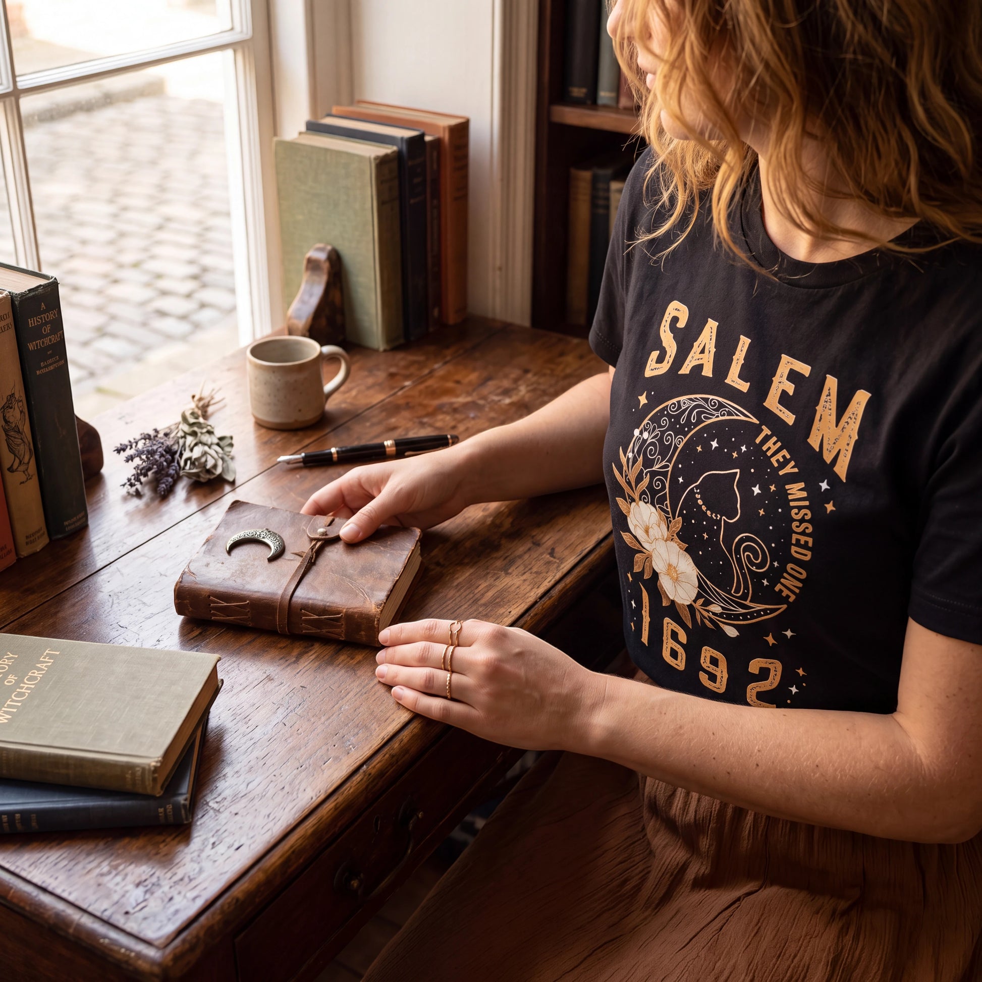 The Witches' Quest black cotton unisex t-shirt featuring a Salem 1692 celestial cat and floral graphic print.