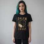 The Witches' Quest black cotton unisex t-shirt featuring a Salem 1692 celestial cat and floral graphic print.