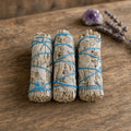 Three 4 inch California white sage smudge sticks tied with blue string on a wooden surface with lavender and crystal